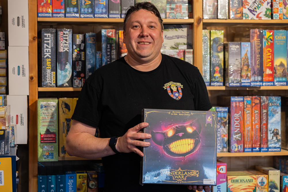 Inhaber Ales hält das Brettspiel 'Wunderland's War' vor einem Regal mit verschiedenen Brettspielen