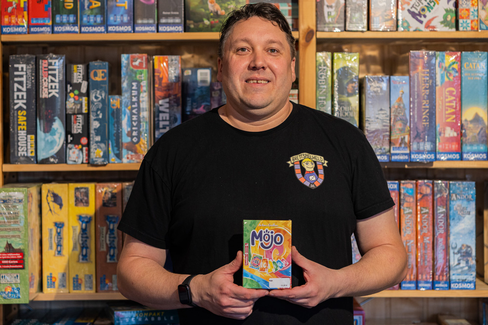 Inhaber Alex trägt schwarzes T-Shirt mit Logo vom Brettspielheld und hält das Kartenspiel 'Mojo' vor einem Regal voller Brettspiele