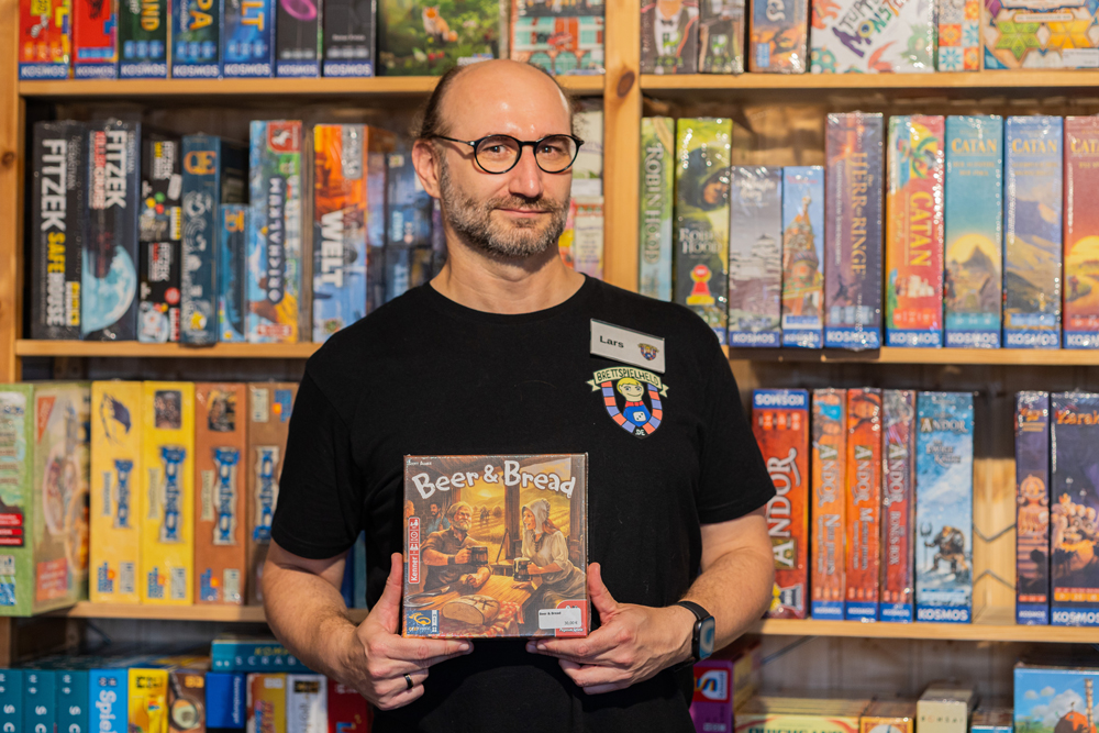 Mitarbeiter Lars hält das Brettspiel 'Beer & Bread' vor einem Regal mit verschiedenen Brettspielen