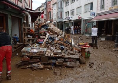 Verschlammte Fußgängerzone mit aufgetürmten beschädigten Brettspielen vor einem Geschäft, Menschen räumen auf