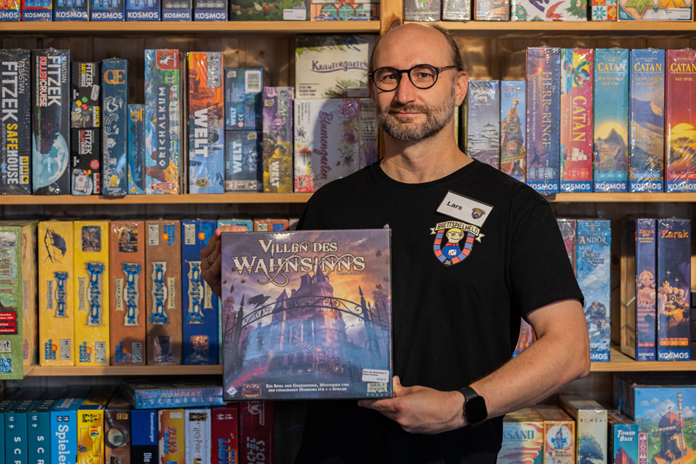 Inhaber Alex trägt schwarzes T-Shirt mit Logo vom Brettspielheld und hält das Kartenspiel 'Mojo' vor einem Regal voller Brettspiele
